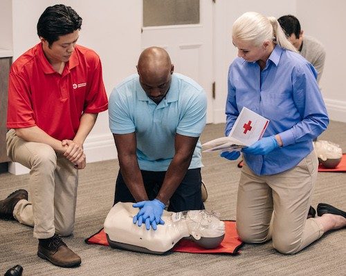 red cross volunteer oversees CPR training as another man practices on a dummy